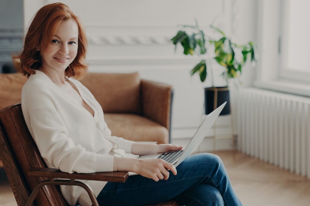 Woman sits in armchair with laptop computer, works remotely, checks email and reads notification